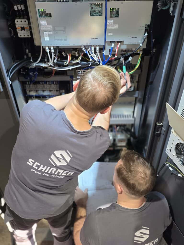 Technicians performing Okuma CNC electronics repair and diagnostic work inside the control cabinet